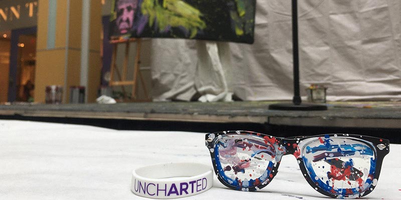 Glasses and a rubber bracelet that says, "Uncharted" in the foreground. The bottom of a canvas with some paint in the background.