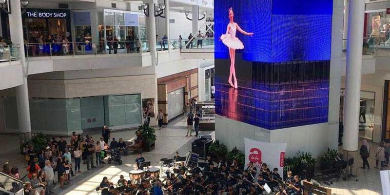 Group of youth orchestra playing in the middle of the mall next to an LED video installation.