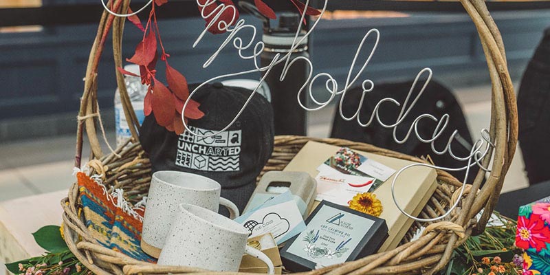 Wicker basket filled with ceramic mugs, soap, hat, and gift cards.